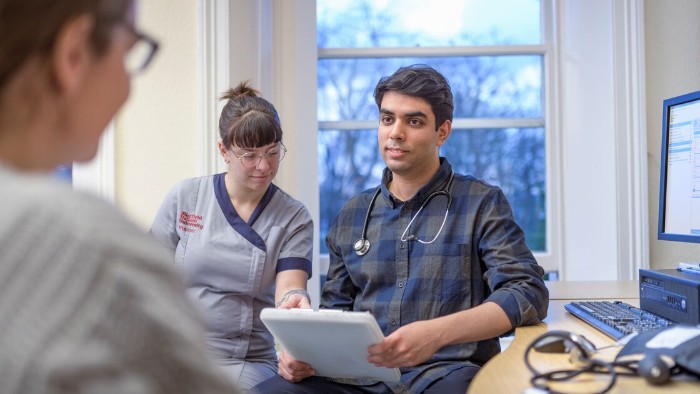 A doctor sat at their desk in discussion with a patient. Next to him is a Sheffield Hallam student in uniform pointing to information on a mobile device.