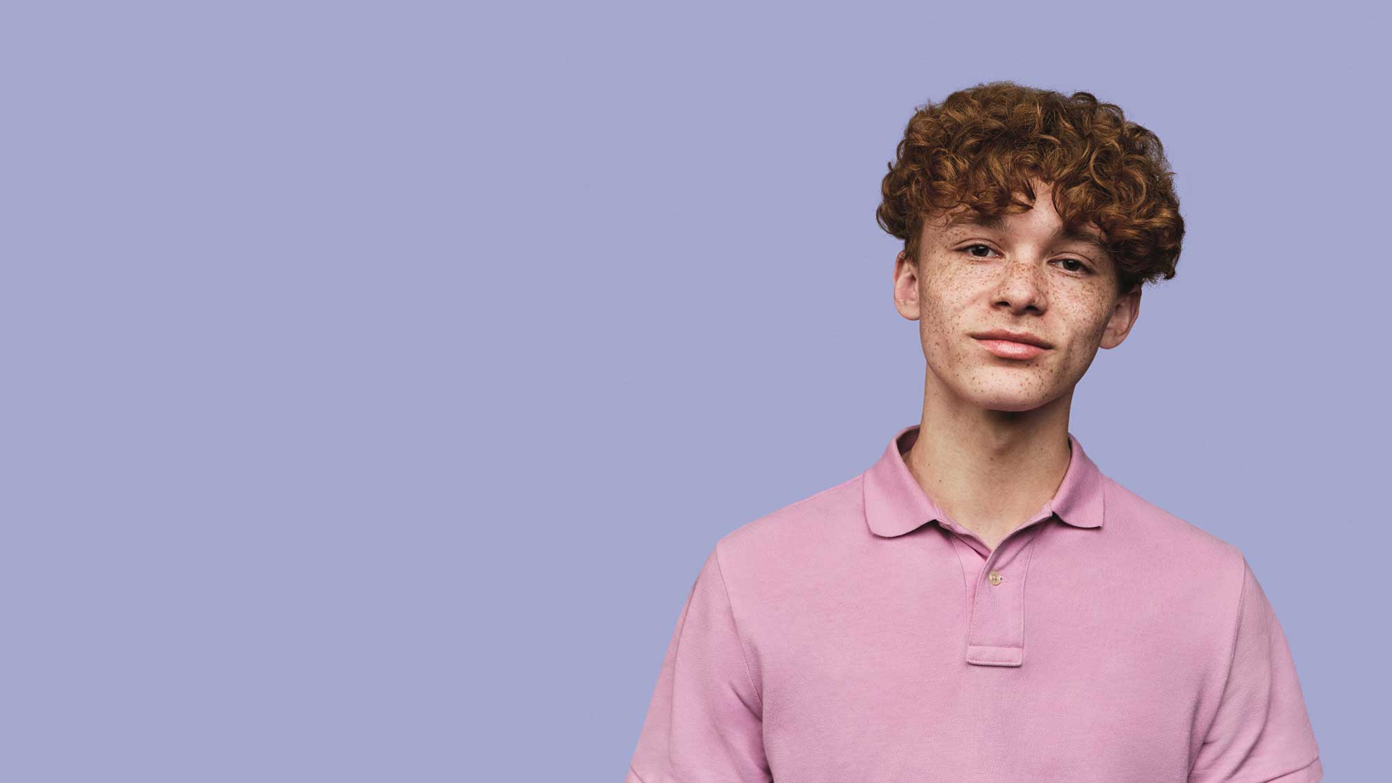 A young male with curly hair and freckles wearing a pink polo shirt, standing against a plain lavender background for a photoshoot.
