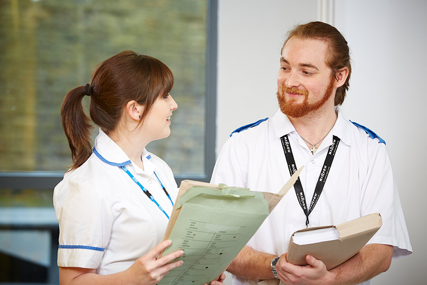 White male with beard smiling at white woman with brown hair holding a folder, both wearing white clinical uniform.