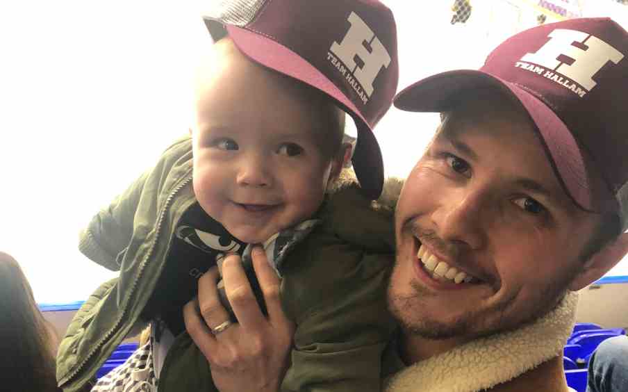 Man holding a baby in the stands of an ice rink with Sheffield Hallam branded caps on.