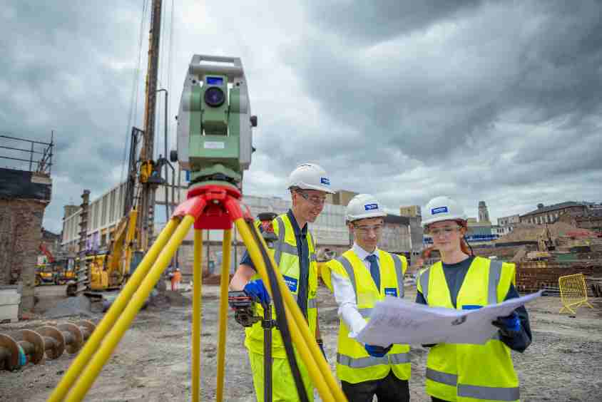 Three people looking at construction plans on a construction site next to quantity surveying equipment.