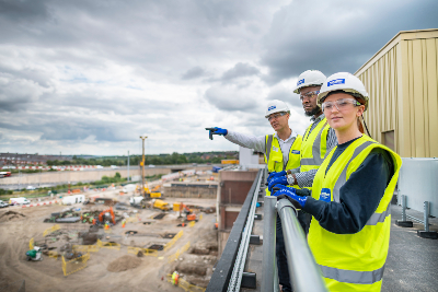 Three people in hard hats and hi-vis vests on the balcony looking over the bare construction site with one of them pointing.