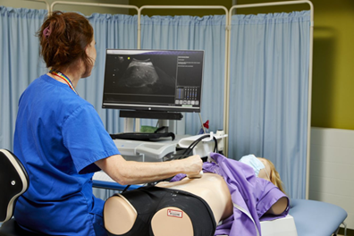 Female student wearing blue scrubs conducts ultrasound on a manakin laid on a bed beside her.