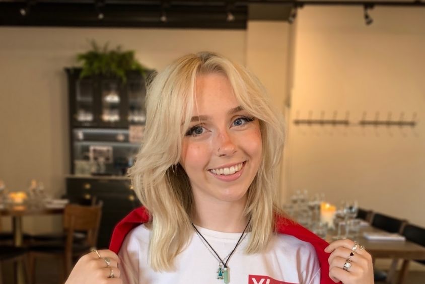 Head and shoulders photo featuring a smiling, blonde haired woman wearing a white t-shirt and red jacket - standing in a restaurant.
