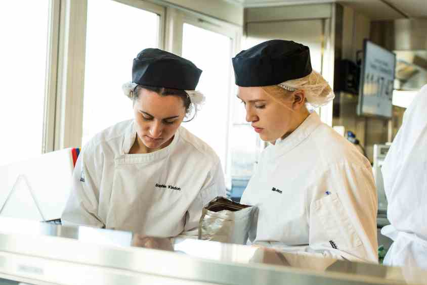 Two women wearing overalls, hair nets and hats in a commercial kitchen while looking down at worktop.