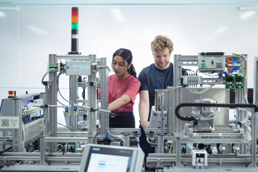 Front facing image of a dark haired female student wearing a dark salmon coloured t-shirt, standing beside a light haired male student wearing a navy t-shirt - both operating complex engineering machinery in front of them whilst stood in a white laboratory environment.