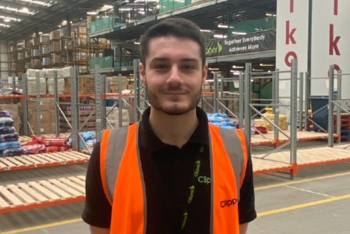 Dark haired male stands in a packaging warehouse wearing a hi-vis vest