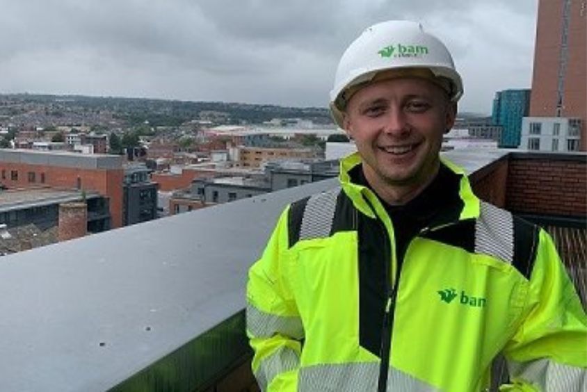 Short haired male smiles to the camera whilst wearing a white hard hat and thick hi visibility jacket. Individual is stood on a rooftop terrace with buildings in the background.