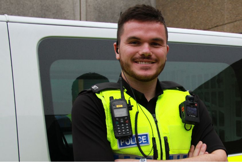 A police officer in uniform smiling stood with arms folded in front of a police van