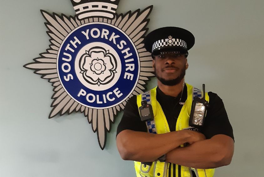 Bearded male wearing a hi-vis police uniform and cap stands before an acrylic shield for South Yorkshire Police.
