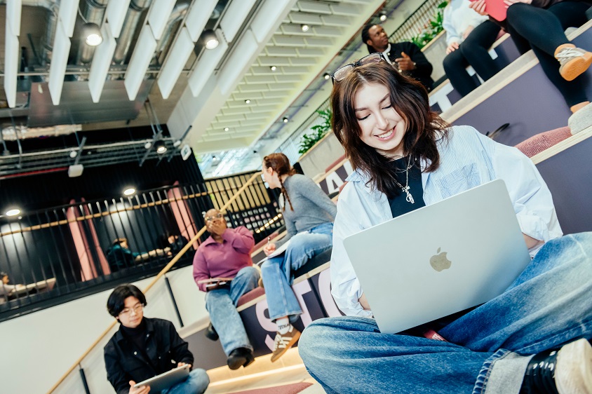 A student sits on tiered seating using a laptop in a modern university study space, while other students work and socialise nearby.