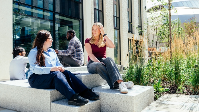 Two students sit on concrete blocks in front of the Howard Street buildings, they are surrounded by the greenery of Hallam Green