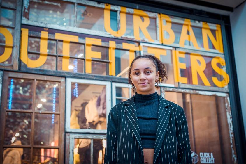 Lauren Riley stands for a photo in front of an Urban Outfitters store with large yellow signage, wearing a black and white pinstripe blazer.