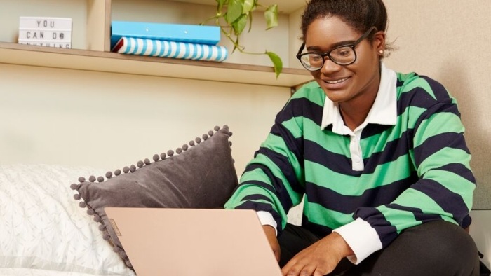 A woman in glasses and a striped top sits on a bed at home using a laptop.