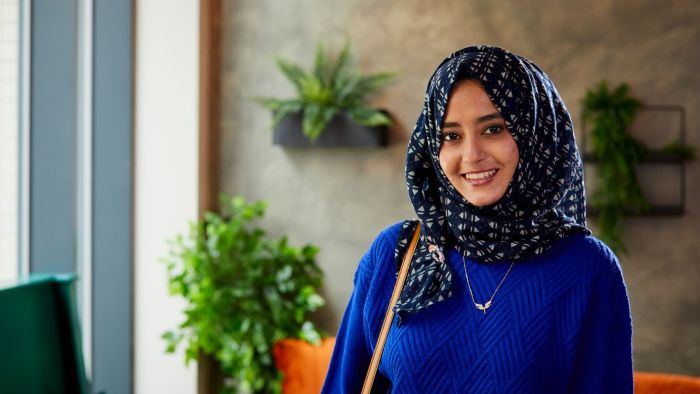 A person wearing a blue sweater and a patterned headscarf stands indoors with decorative plants in the background.