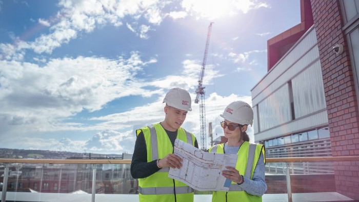 Two students on the terrace of a building wearing hard hats and high visibility jackets. Both are looking at a detailed paper plan.
