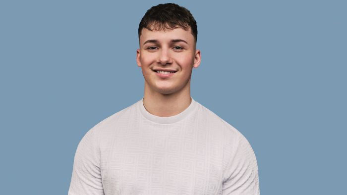 A young man wearing a white textured shirt stands against a solid blue background smiling for the camera.