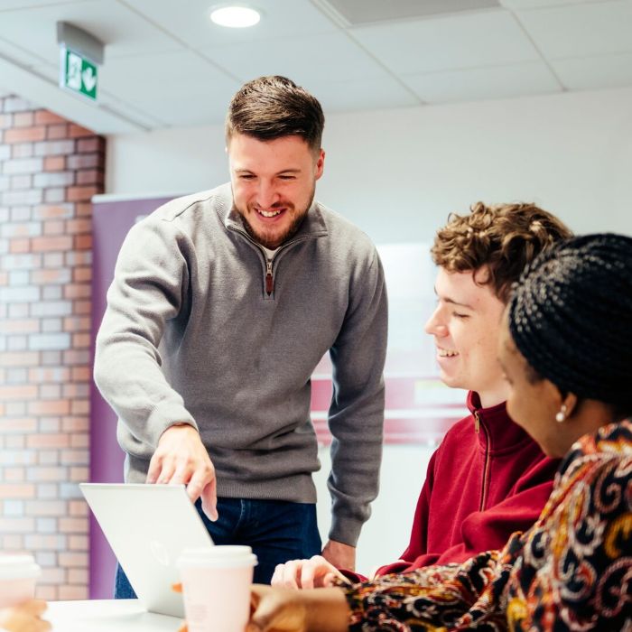 A person in a grey sweater points at a laptop screen in use by two others, both sat at a table on campus.
