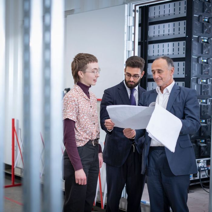 Three smartly dressed people stand in a server room looking at documents, with a large server rack behind them.