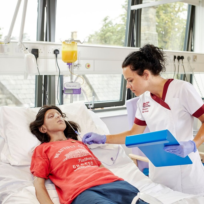 A nursing student in uniform cares for a patient in a simulated hospital ward, holding a clipboard beside a hospital bed.