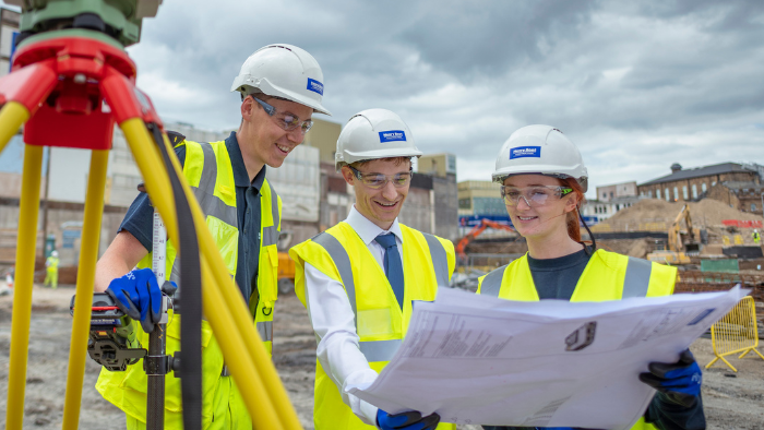 Three young construction and surveying workers stand together in the centre of a site beside surveying equipment whilst wearing gloves, hard hats and hi-vis jackets as they review a site plan together.