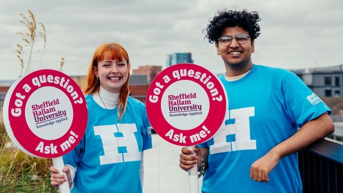 Two open day ambassadors in blue Hallam t-shirts stand on the rooftop of a new building, smiling and holding signs saying 'Got a question? Ask me!'