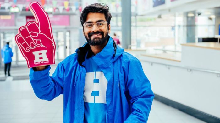 A smiling open day ambassador in a blue jacket holds a large foam finger with "Team Hallam" written on it, he is stood in the entrance to the City Campus.