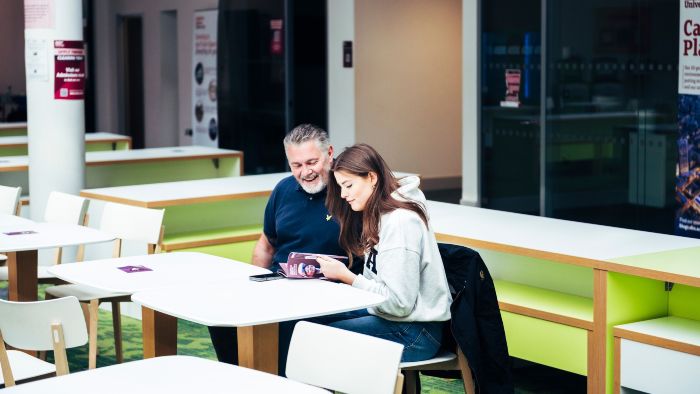 A father and daughter sat at a table in a campus dining area, with the girl holding open a booklet they are reading through together.