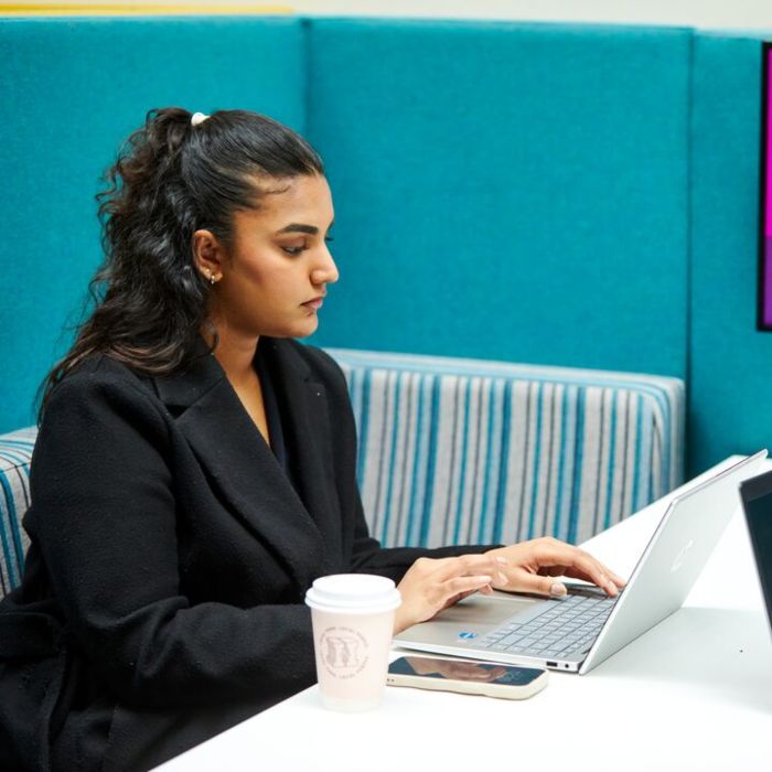 A person in a smart black coat sits at a table on campus using a laptop, with a coffee cup and smartphone nearby.