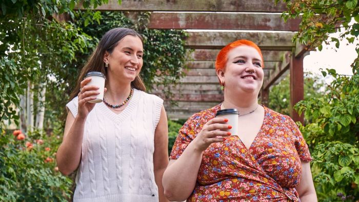 Two smiling people walk outdoors under a wooden pergola, each holding a takeaway coffee cup, surrounded by greenery.