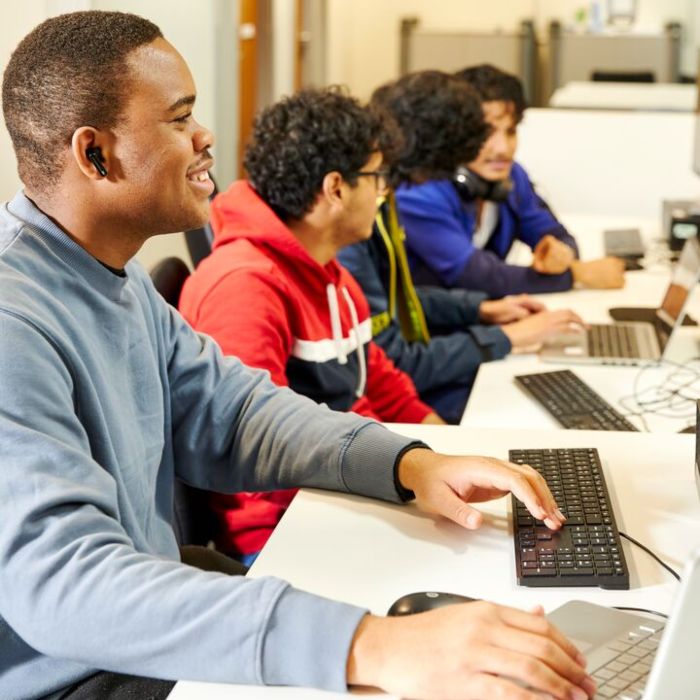 Four students sat alongside each other at a long classroom table. They are using desktop and laptop computers, working and studying together.