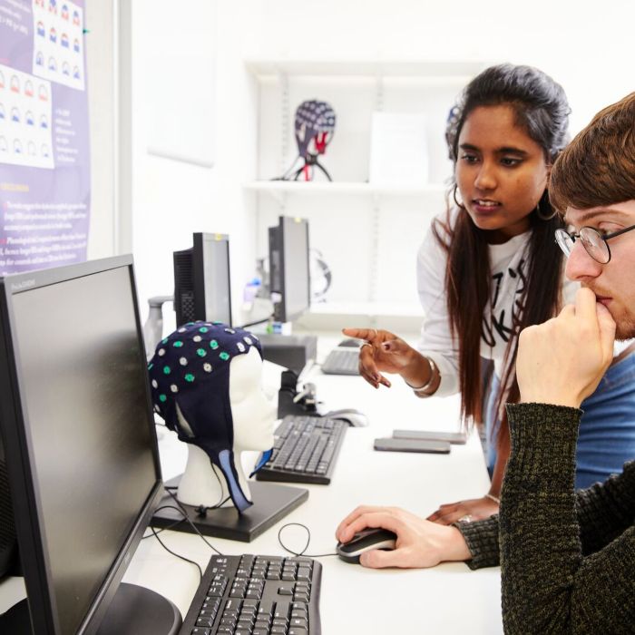 Two students in a classroom, one using a computer and the other pointing at the screen. Nearby is a mannequin head wearing an EEG cap on the desk.