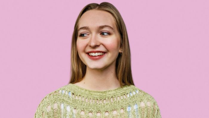 Against a pink background, a young woman with long blonde hair and green top is looking to the left and smiling.