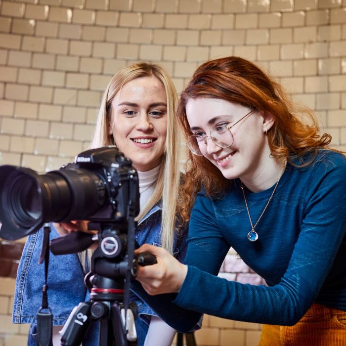 Two female students are smiling while they set up and look through a camera on a tripod.