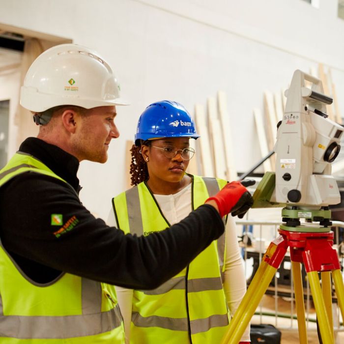 A man and young woman are on a construction site in high vis jackets and hardhats, setting up surveyor equipment on a tripod.