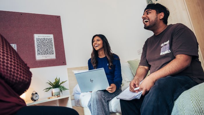 Three students sitting in a halls bedroom, talking and laughing while using a laptop and notebook.
