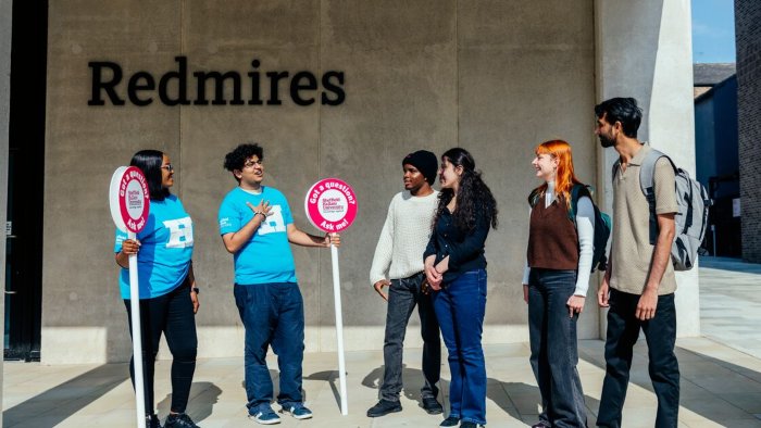 A group of people standing outside the Redmires campus building, talking with two student open day ambassadors holding signs.