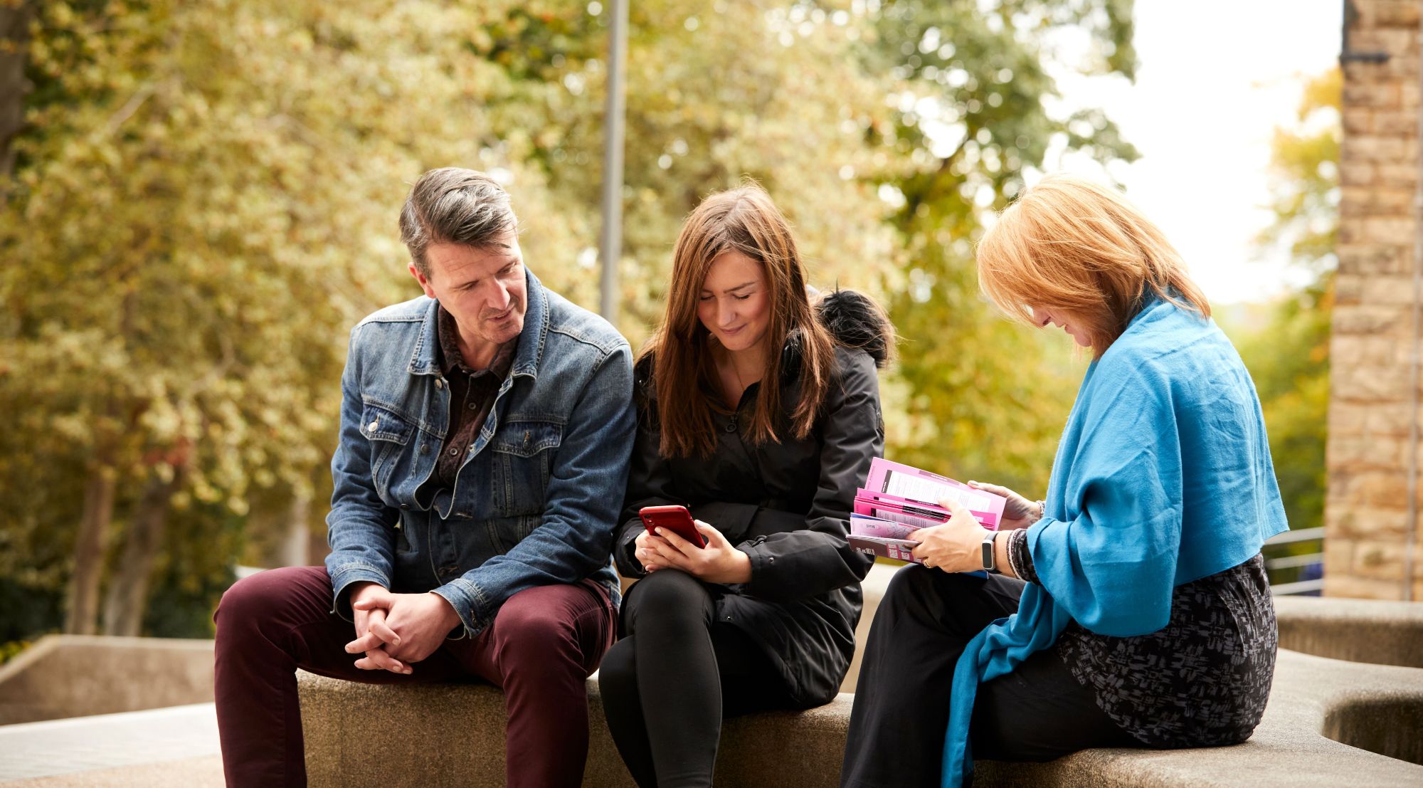 Two parents sat with their daughter on an outdoor bench at Heart of the Campus, looking at a mobile phone and a printed open day guide.