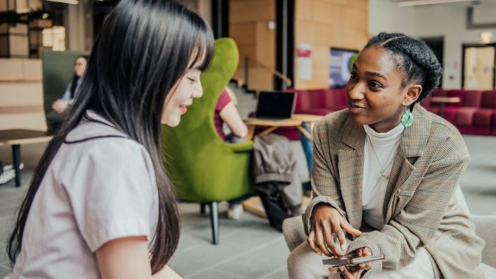 Two students sat talking in a modern lounge area at Sheffield Hallam campus while another person works on a laptop nearby.