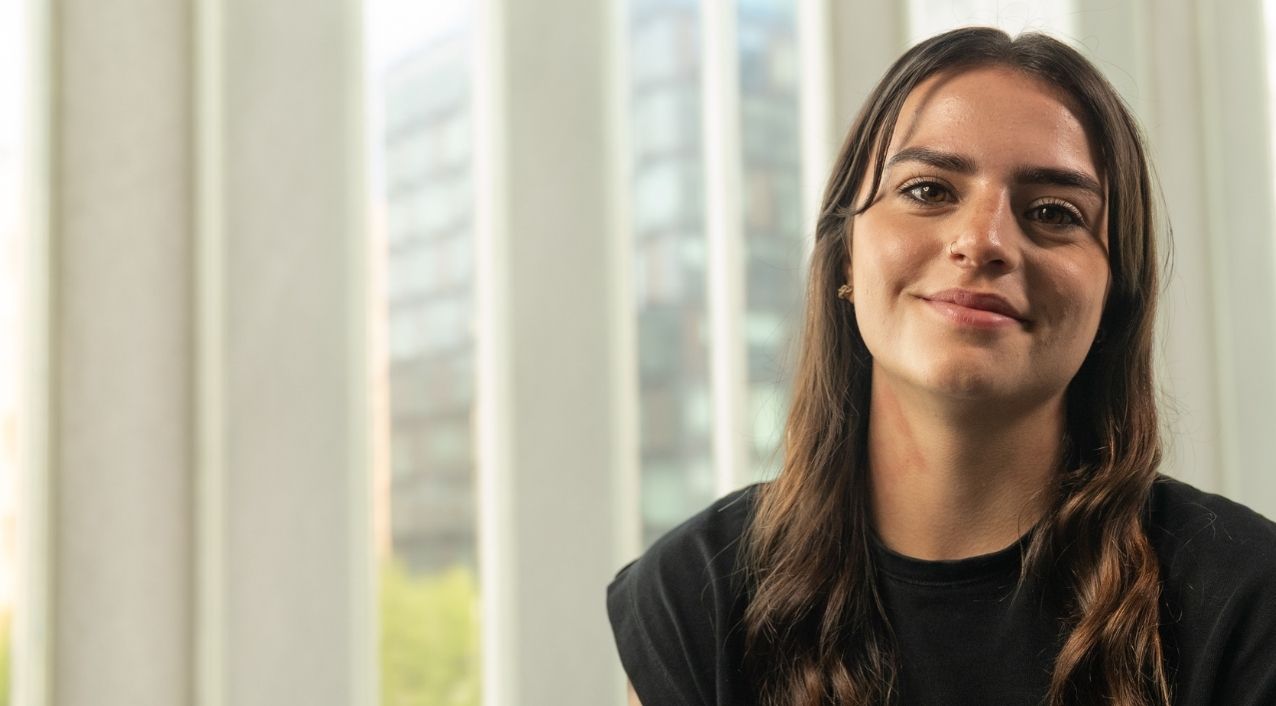 Leah is smiling, has long brown hair and is wearing a black t-shirt. There is a window in the background with a blurred view of a large office building.