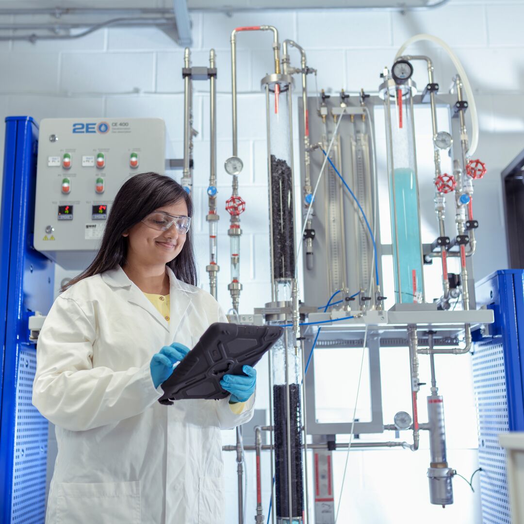 A woman in a lab coat wearing protective equipment holds a tablet, engaged in her work in a laboratory setting.
