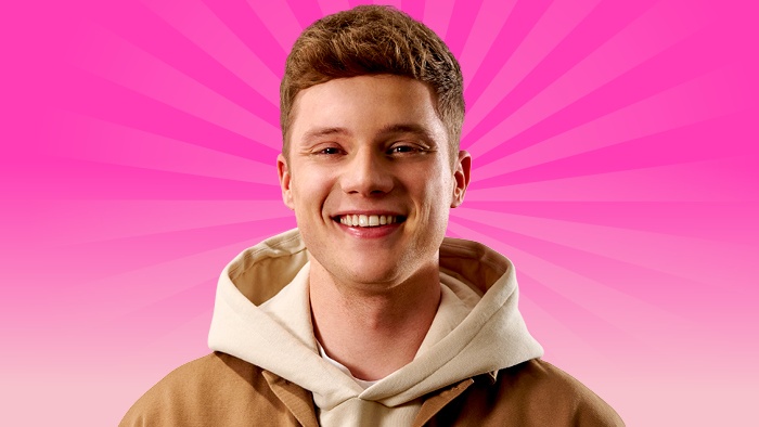 A young male student in a brown jacket and hoodie standing against a bright pink, radiating background.