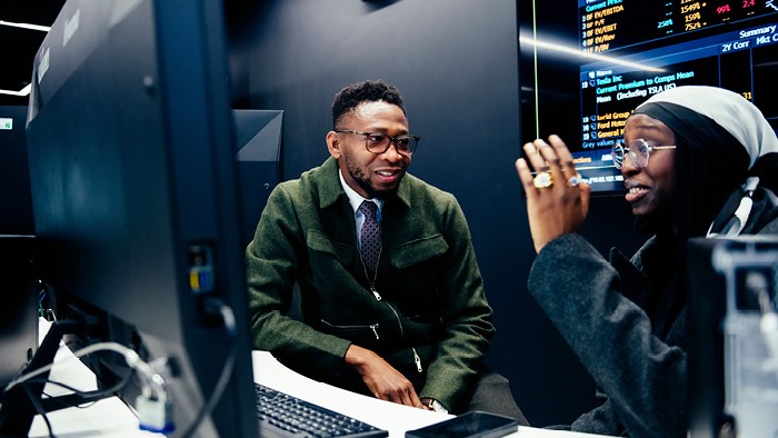 Two students sat talking at a desk in front of a PC monitor. Behind them is a big screen showing financial data.