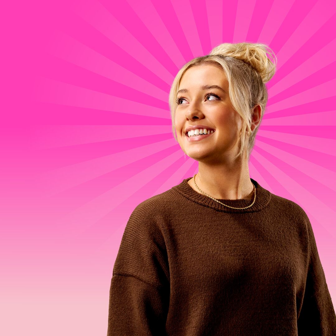 A female student with blonde hair and wearing a brown jumper, standing in front of a bright pink radiating background.