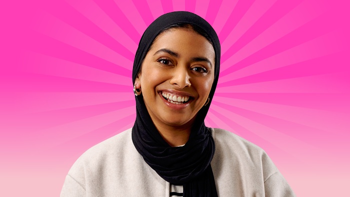 A female student wearing a black headscarf and light jacket, smiling in front of a bright pink radiating background.