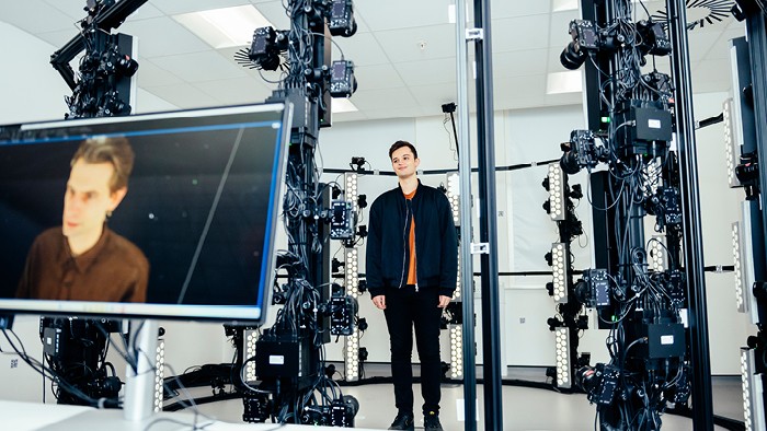 A male student standing inside a multi‑camera 3D scanning rig, with a monitor in front showing his digital scan.