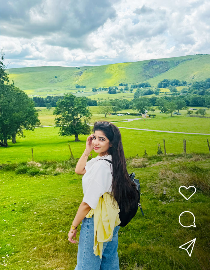 A woman standing in a green countryside landscape, looking back toward the camera with hills, trees and cloudy skies in the background.