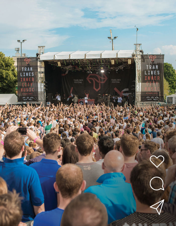 A large crowd gathered outdoors in front of a stage at the Tramlines music festival, with performers on stage and banners displaying the festival name.