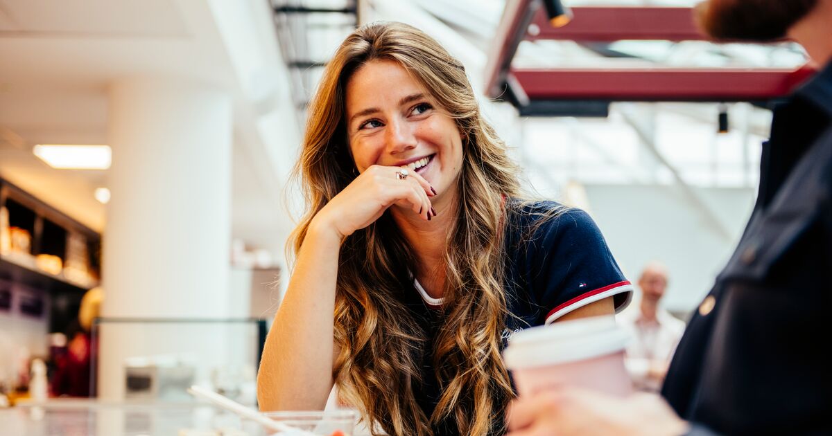 A person with long hair sat at a table in Hallam Cafe, smiling at another person holding a hot drink next to them 