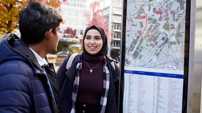 Two people stood looking at a large streetside map of Sheffield city centre. In the background are trees and modern office buildings.